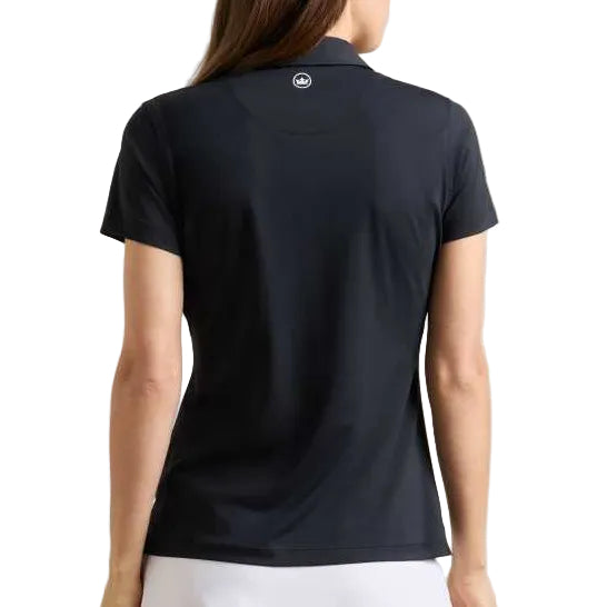 A woman shown from the back wearing a dark navy short-sleeve polo shirt and a white tennis skirt against a plain light beige studio background.