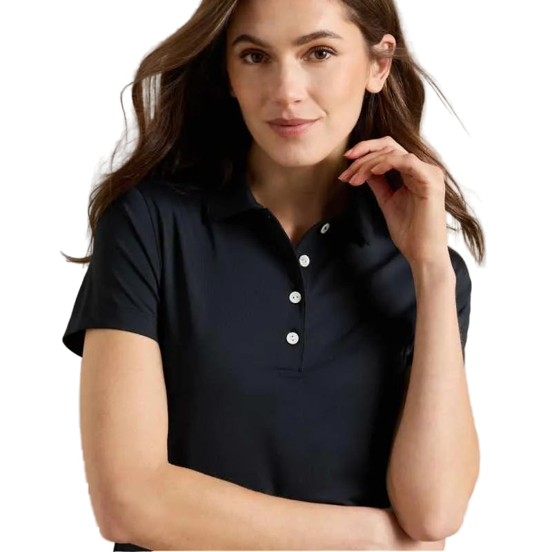 A woman wearing a dark navy short-sleeve polo shirt with white buttons, posing against a light gray wall.