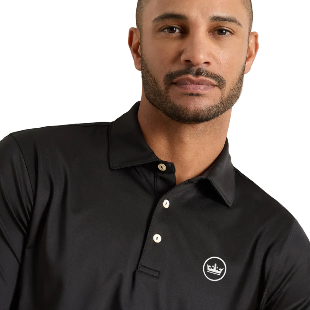 A man wearing a black long-sleeve polo shirt with a small white embroidered crown logo on the left chest, photographed against a plain light gray wall.