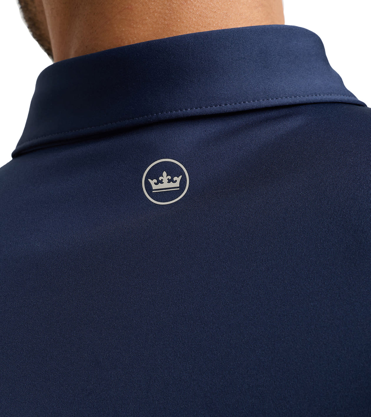 Close-up of the back of a navy blue polo shirt on a model, featuring a small white embroidered crown logo inside a circle on the upper back.