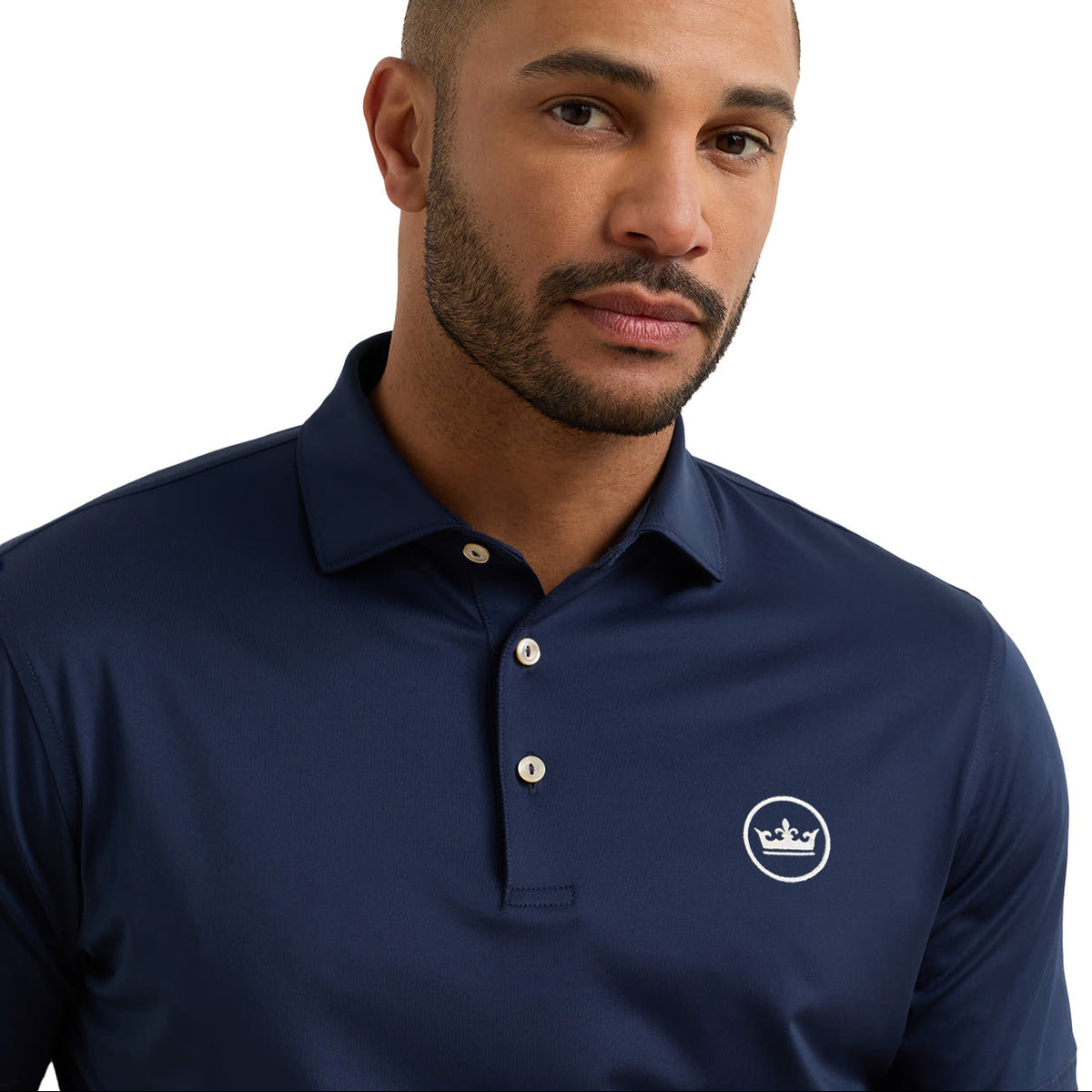 A man wearing a navy blue polo shirt with a small white embroidered crown logo on the left chest, photographed against a clean white background.