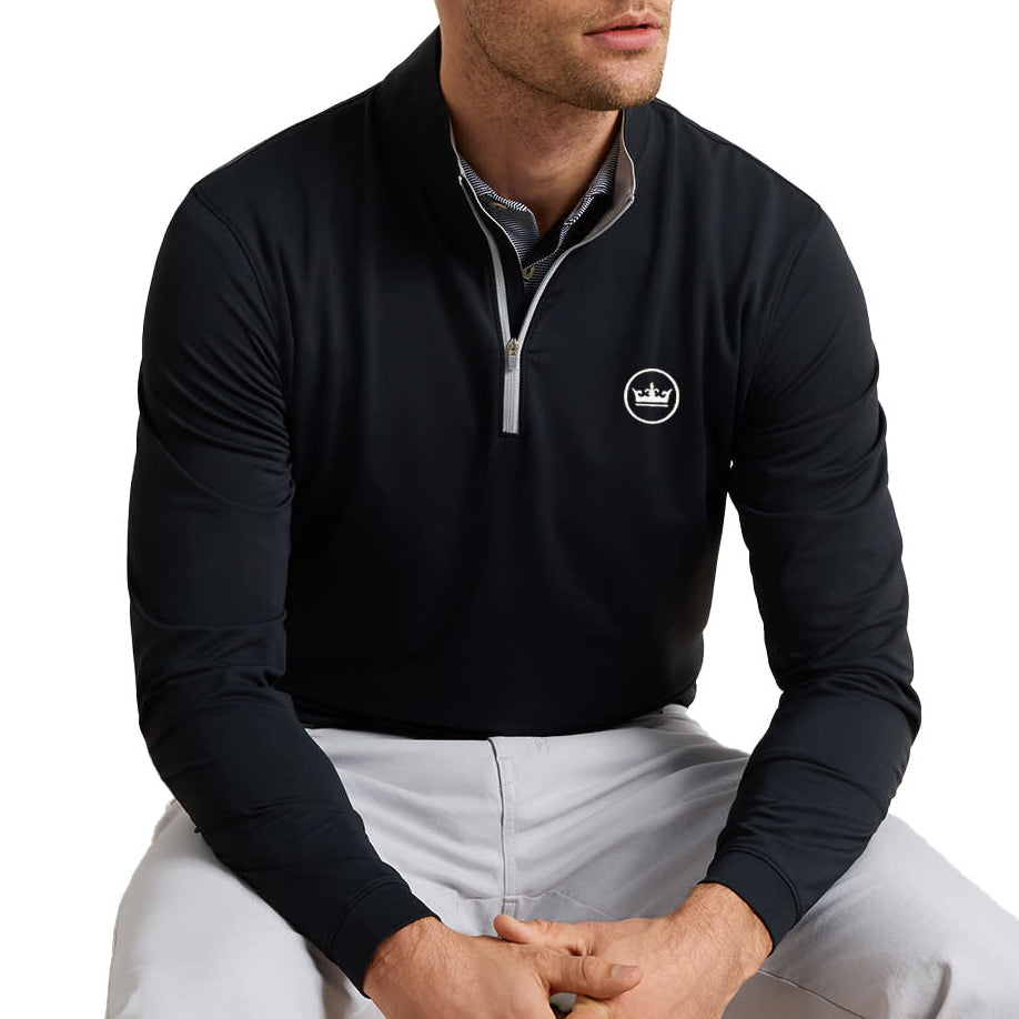 A man wearing a black long-sleeve quarter-zip pullover with a white circular logo on the left chest, paired with light gray pants, seated against a solid white background.
