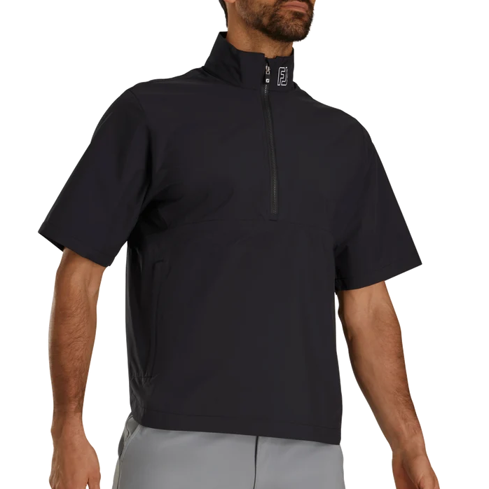 A man wearing a black short-sleeve quarter-zip pullover top with a high collar and gray pants, shown from the waist up against a plain white background.