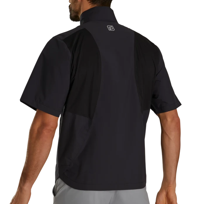 Rear view of a man wearing a black short-sleeve athletic shirt with a high collar and a small white logo on the upper back, paired with light gray pants, against a plain white background.