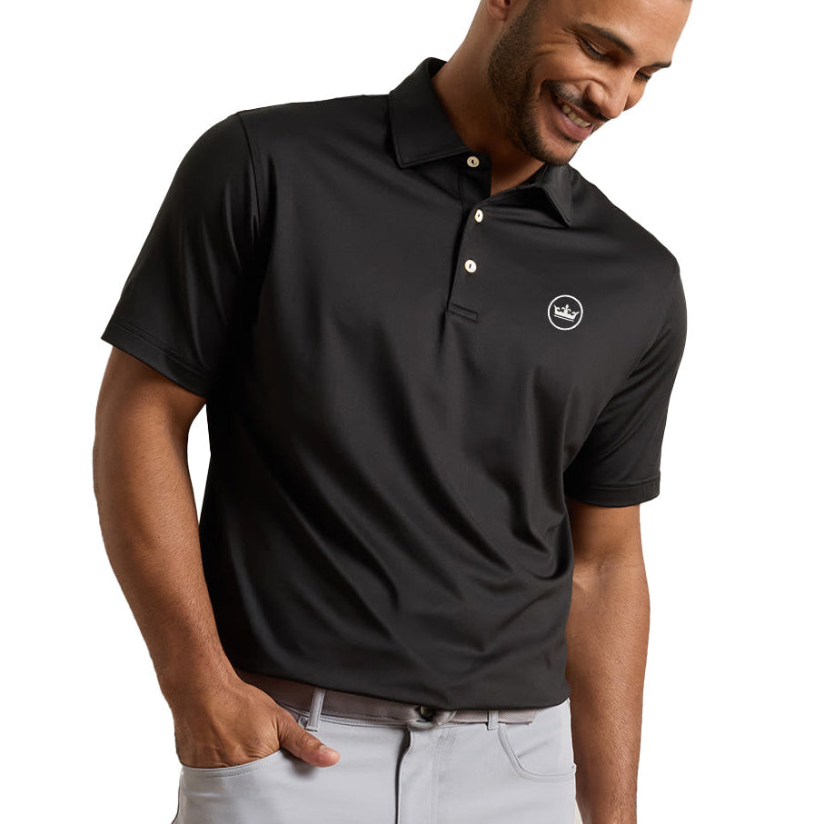 A man wearing a black short-sleeve polo shirt with a small circular logo on the left chest, paired with light gray pants, photographed against a plain white background.