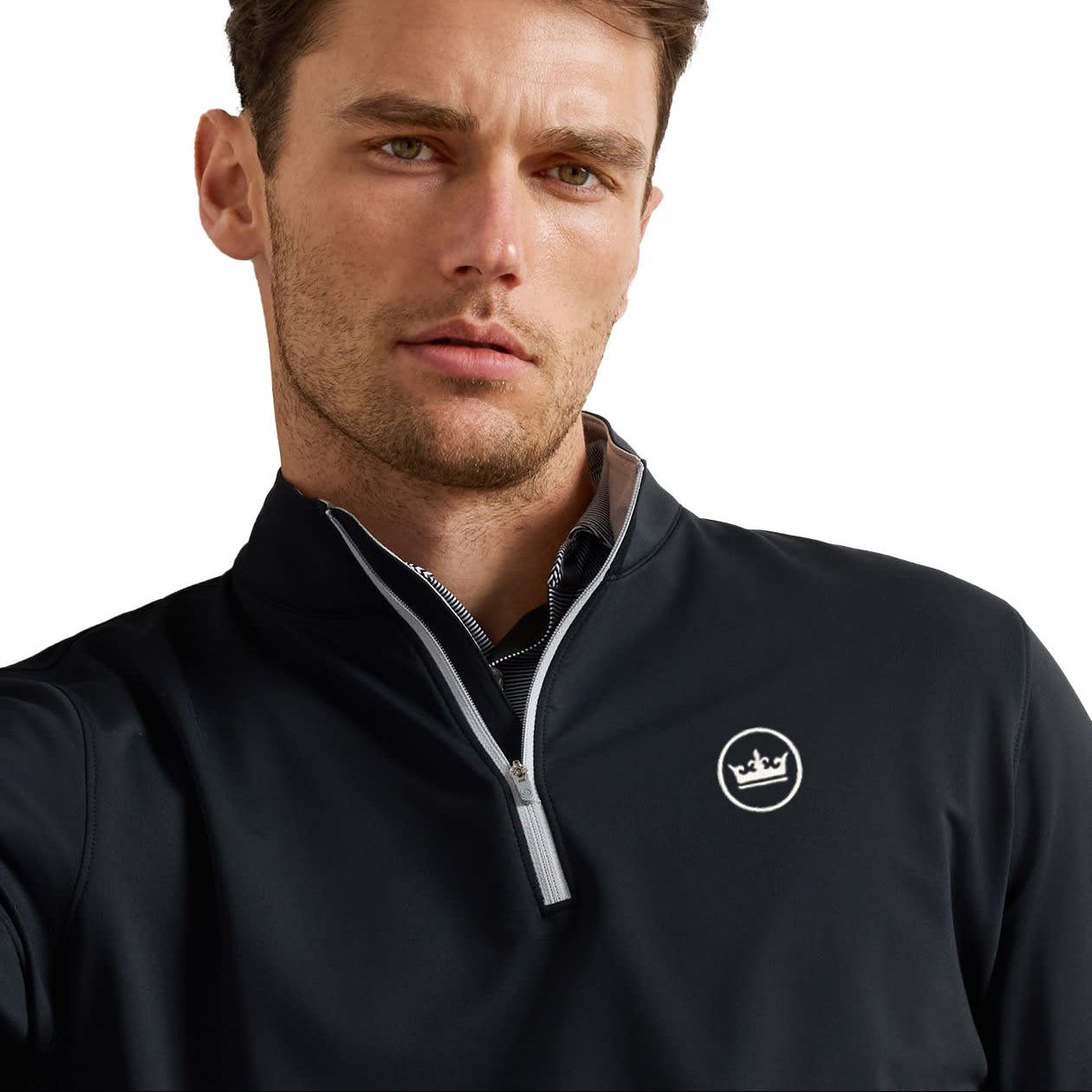 A close-up of a man wearing a dark navy quarter-zip pullover with a white circular logo featuring a crown on the left chest, set against a plain white background.